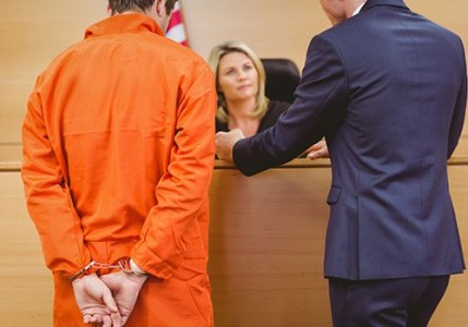 A prisoner wearing an orange jumpsuit is standing next to his lawyer, talking to a judge at her bench.