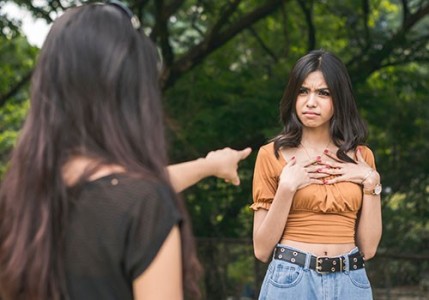 A teen girl is pointing at another teen girl who's body language is saying "me?"