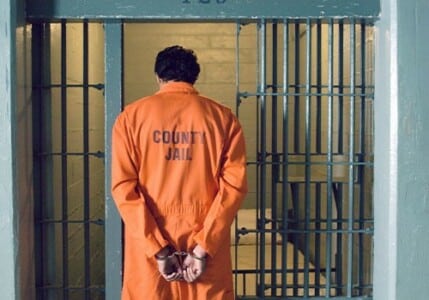 An inmate in an orange jumpsuit and handcuffs walking into his prison cell.